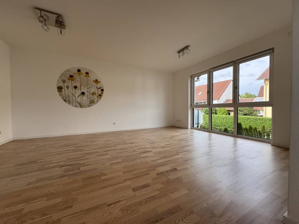 Leerer Raum mit Holzboden und großen Fenstern, mit Sicht auf eine Hecke
