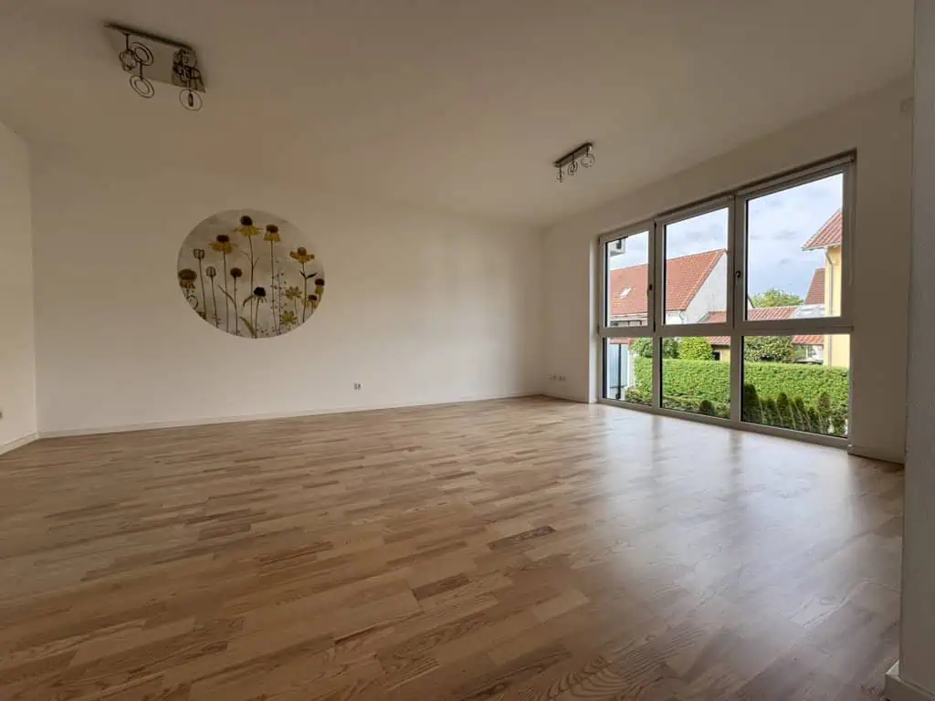 Leerer Raum mit Holzboden und großen Fenstern, mit Sicht auf eine Hecke